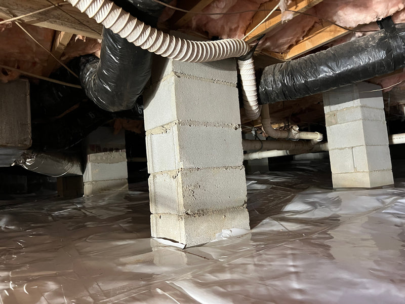 Concrete block pier in a crawlspace with HVAC flex ducts and PVC pipes routed overhead above a partially installed vapor barrier