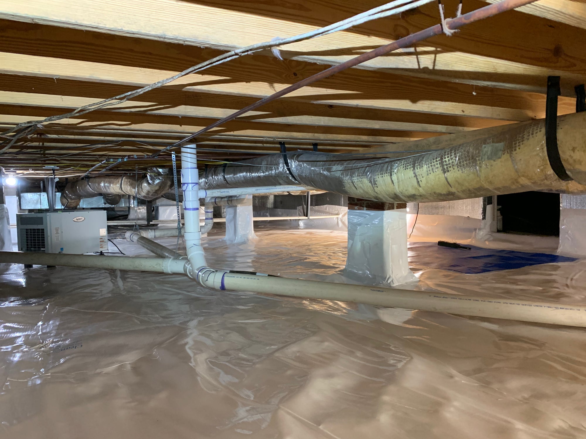 Clean crawlspace interior with a vapor barrier covering the ground, HVAC ducts running overhead, and a dehumidifier unit visible in the background