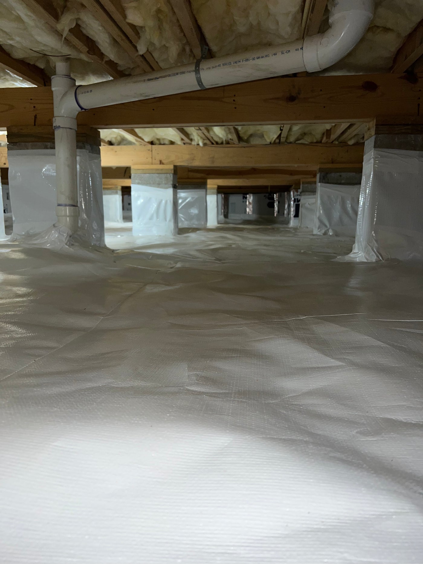 Wide crawlspace with a full vapor barrier encapsulation covering the floor and piers, with PVC plumbing running along the ceiling joists