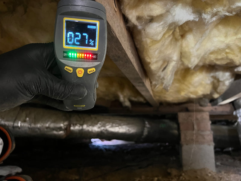 Wood moisture meter reading 27 percent held near deteriorating insulation and plumbing in a crawlspace