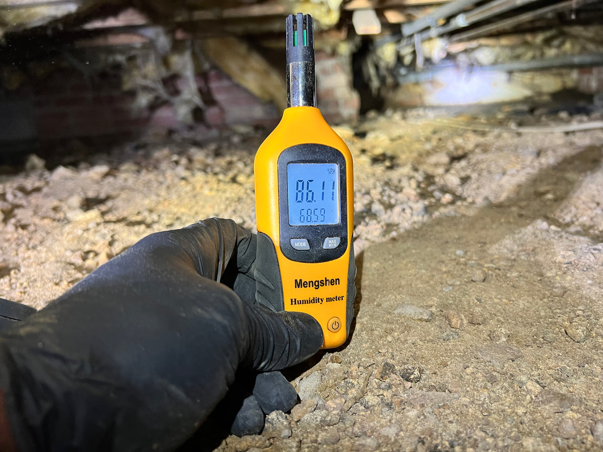 Technician holding a humidity meter reading 86% inside a crawlspace with deteriorated materials visible