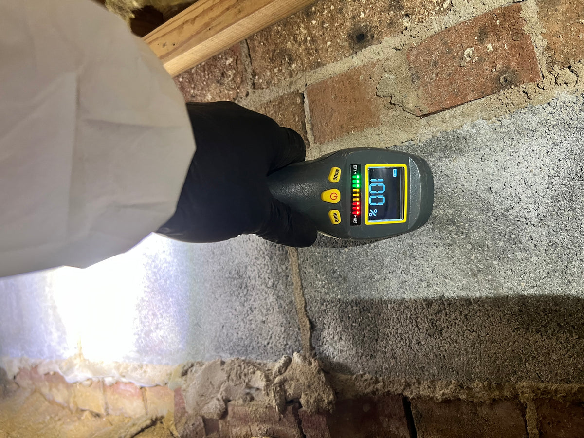 Moisture meter held against a crawlspace foundation wall reading 100 percent saturation